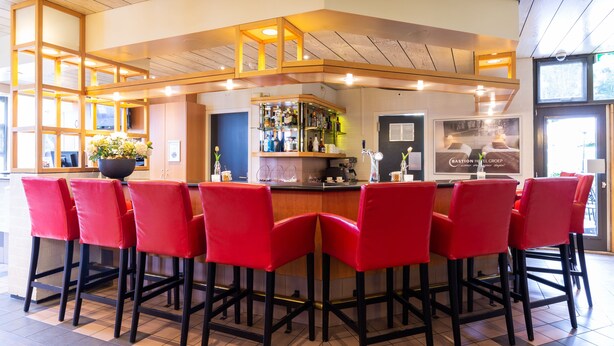 A bar area with red bar stools, a wooden ceiling, and a visible sign for the Easton Hotel Group. - Bastion Hotel Heerlen (Heerlen)