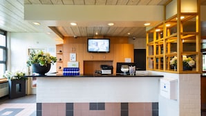 A hotel reception area with a wooden ceiling, a flat-screen TV mounted on the wall, a reception desk, and a potted plant. - Bastion Hotel Heerlen (Heerlen)