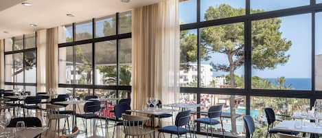Une salle à manger spacieuse, dotée de grandes fenêtres offrant une vue sur la mer et les bâtiments. Les tables sont disposées avec soin et accompagnées de chaises, et le sol est carrelé.