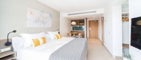 A modern hotel room with a large bed, a desk, and a kitchenette in the background.