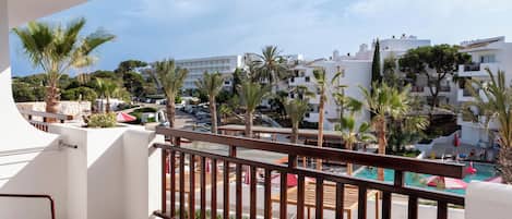 Ein Balkon mit Blick auf einen Pool und Palmen, ein Tisch mit einem Glas sowie ein Bett mit gestreifter Bettwäsche.
