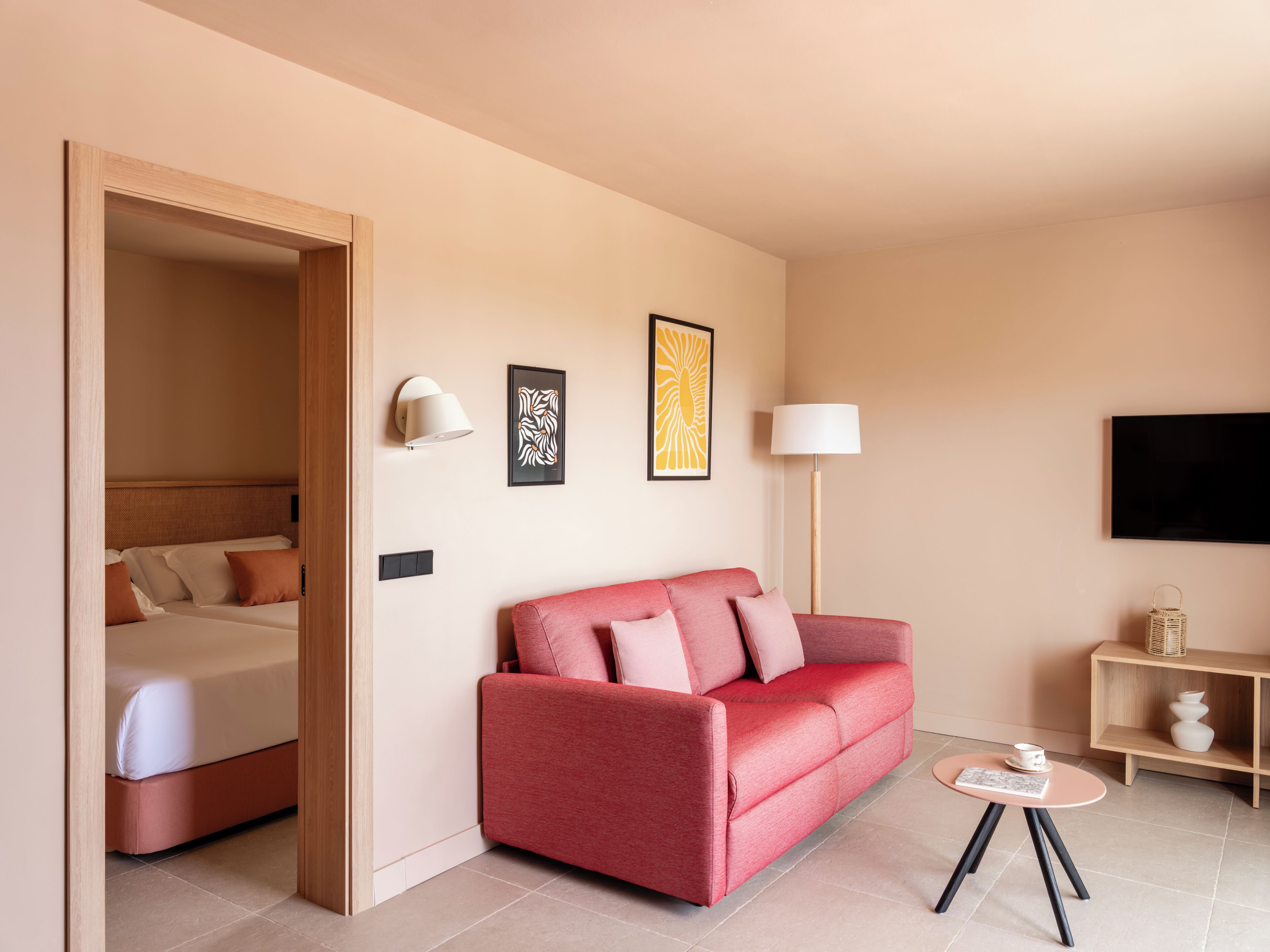 Une chambre d’hôtel avec un lit, un canapé rouge, une petite table, une lampe et des œuvres d’art accrochées au mur.