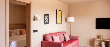 A hotel room with a bed, a red sofa, a small table, a lamp, and artwork on the wall.