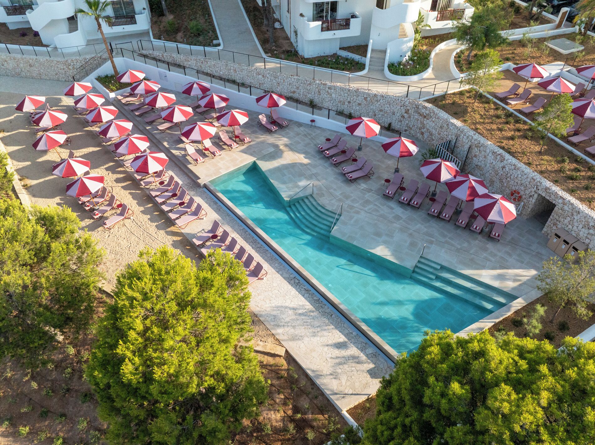 Zona de piscina en la azotea con múltiples tumbonas y sombrillas, rodeada de muros de piedra y vegetación.