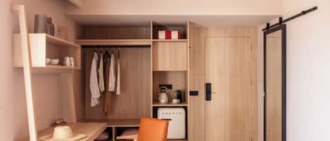A compact room with a wooden desk, a chair, a wardrobe, and a compact kitchenette.