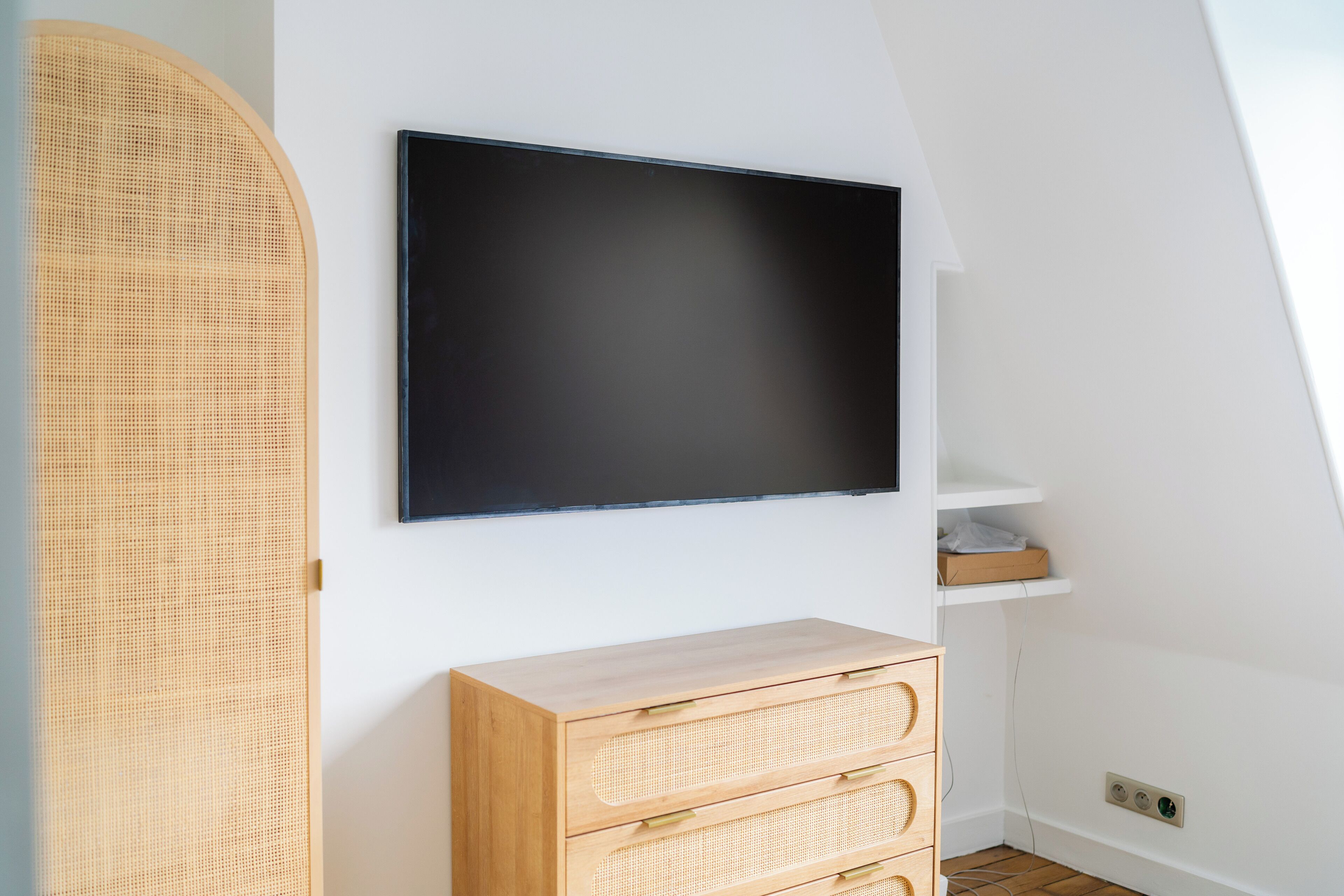 Ein Zimmer mit einem an der Wand befestigten Flachbildfernseher, einer Holzkommode mit Schubladen und einem Korbwäschesammler.