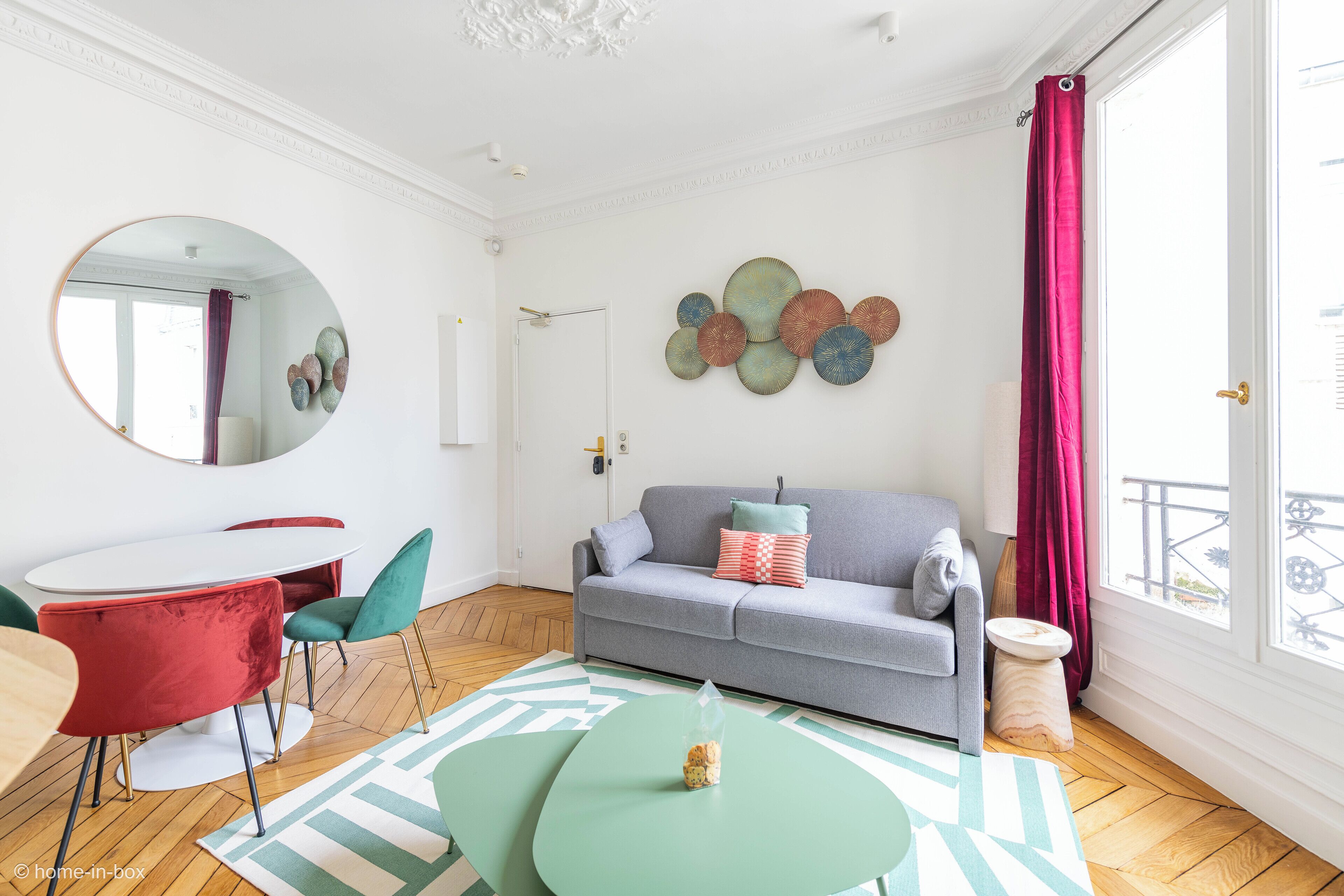 Un salon moderne avec un canapé gris, des fauteuils rouges et turquoise, une table à manger blanche et une grande fenêtre donnant sur un beau paysage.