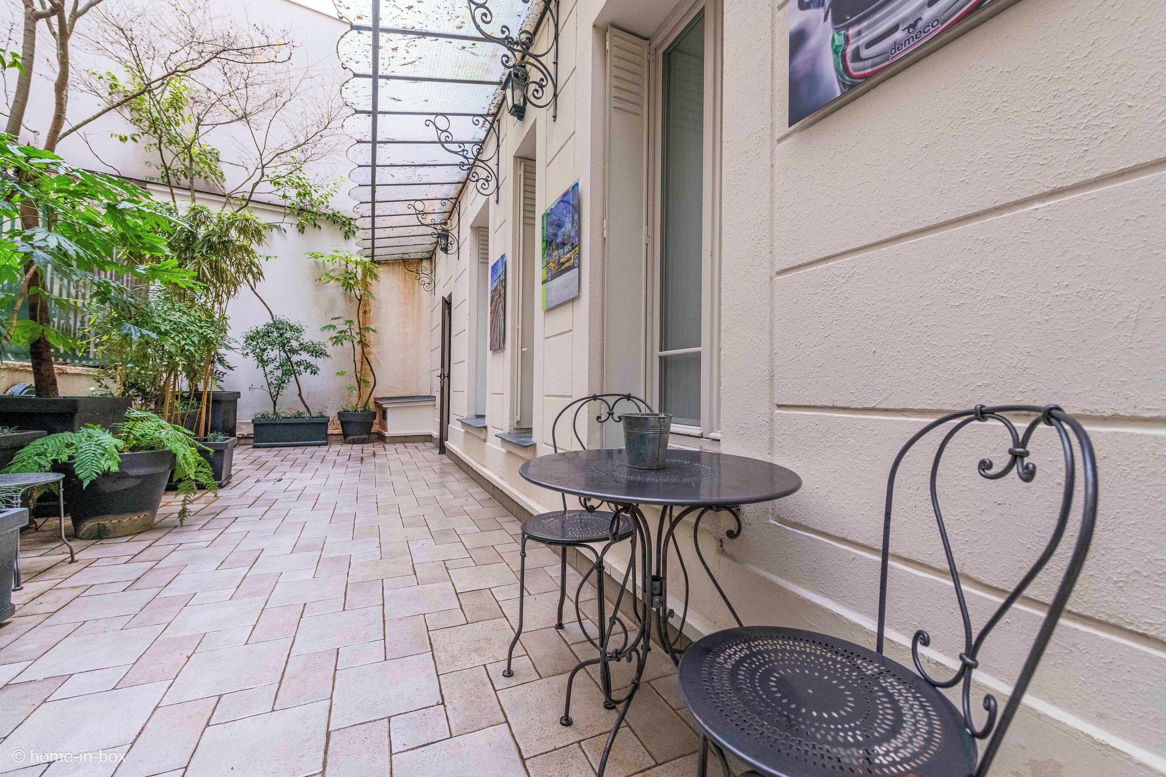 En overdækket terrasse med flisegulv, metalstole og krukker med planter.