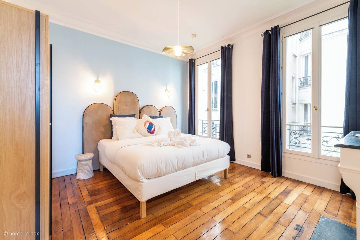 Ein Schlafzimmer mit Holzboden, einem weißen Bett mit Kopfteil, einem Nachttisch und einem Fenster mit Vorhängen.