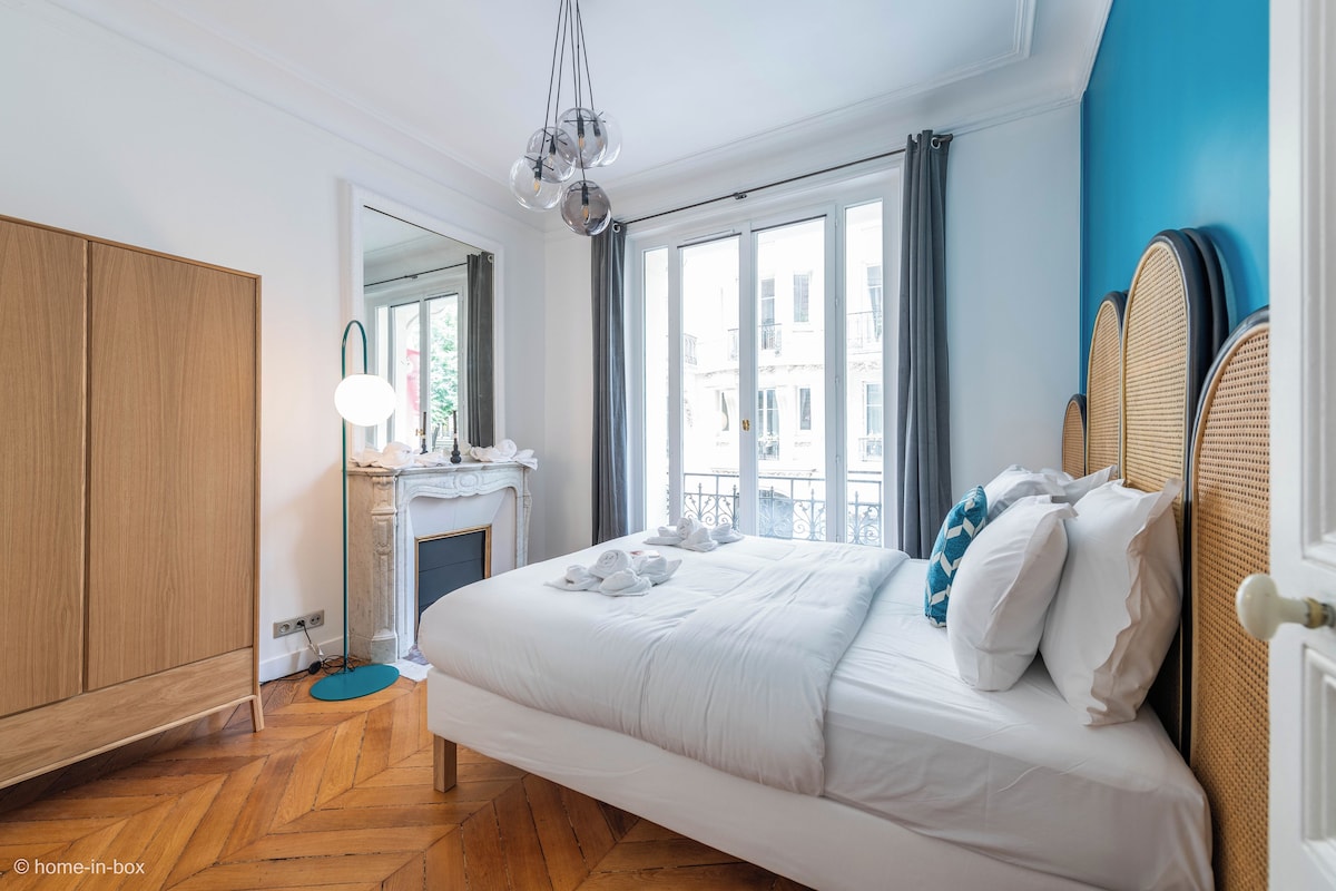 A bedroom with a large bed, a wooden wardrobe, a mirror, and a view of the city through large windows.