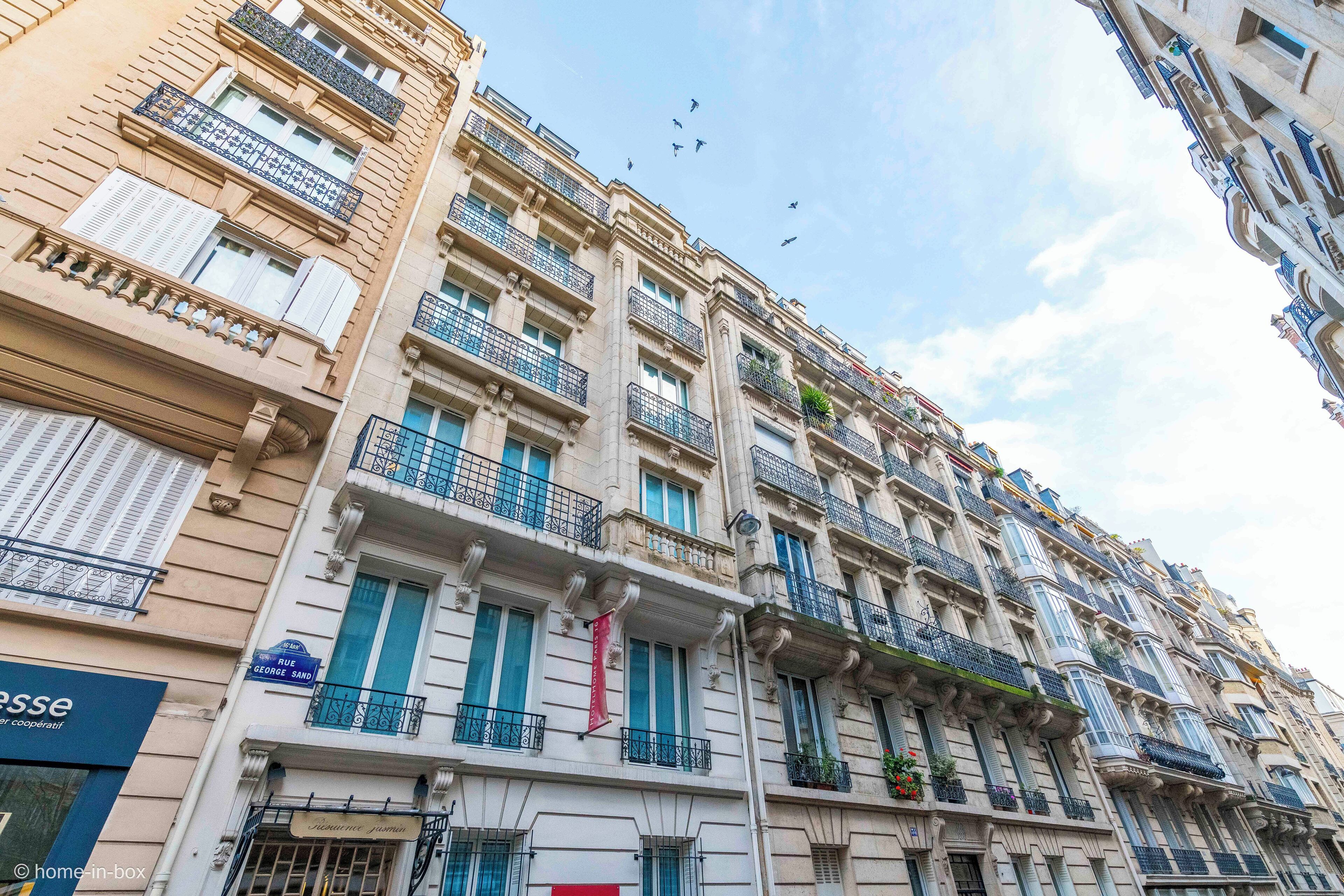 Un immeuble à plusieurs étages doté de balcons, avec un magasin visible au rez-de-chaussée, et un ciel dégagé avec des oiseaux en arrière-plan.