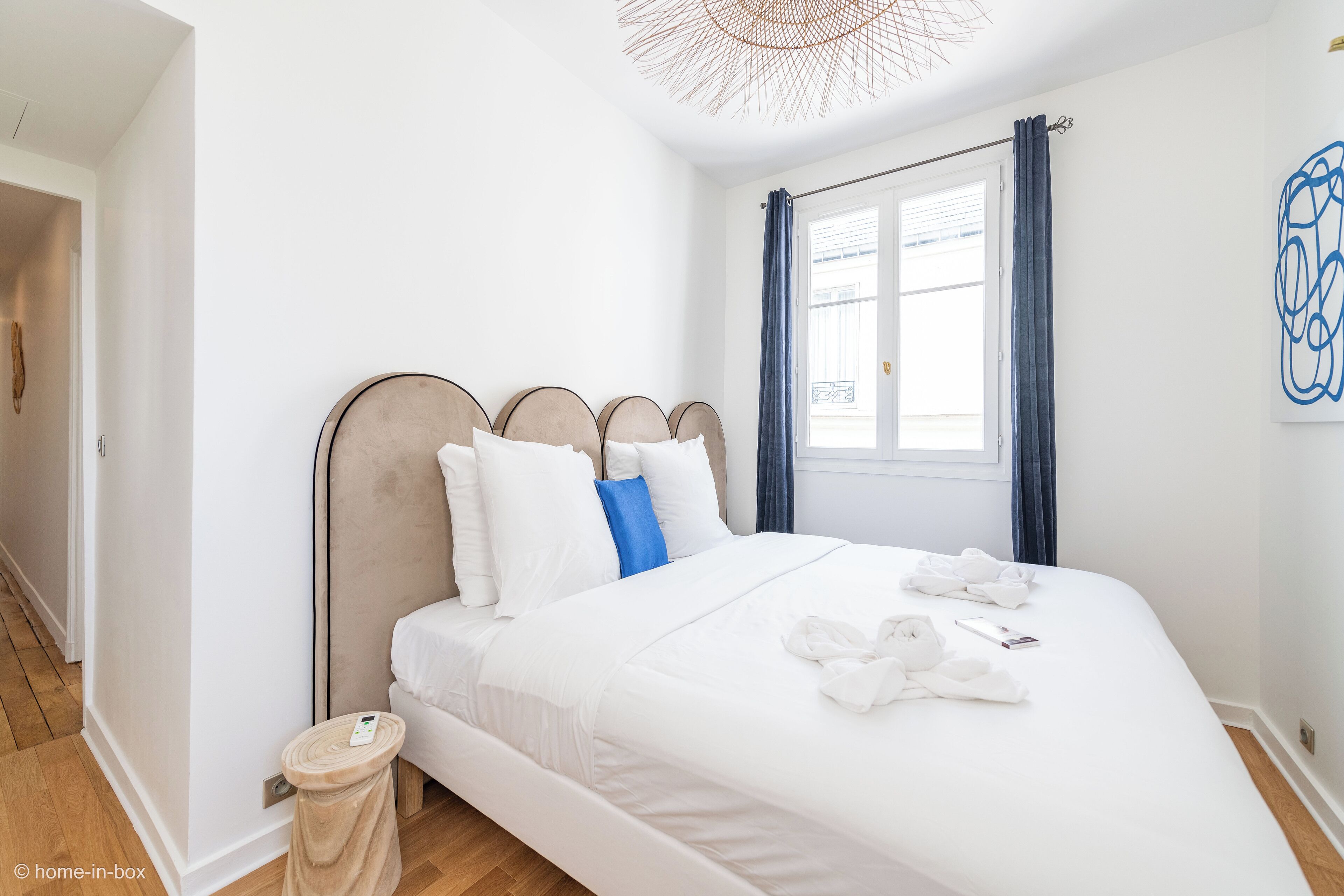 Een netjes opgemaakt bed met witte lakens en een blauw kussen, een nachtkastje met een afstandsbediening, en een raam met blauwe gordijnen.