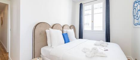 Een netjes opgemaakt bed met witte lakens en een blauw kussen, een nachtkastje met een afstandsbediening, en een raam met blauwe gordijnen.