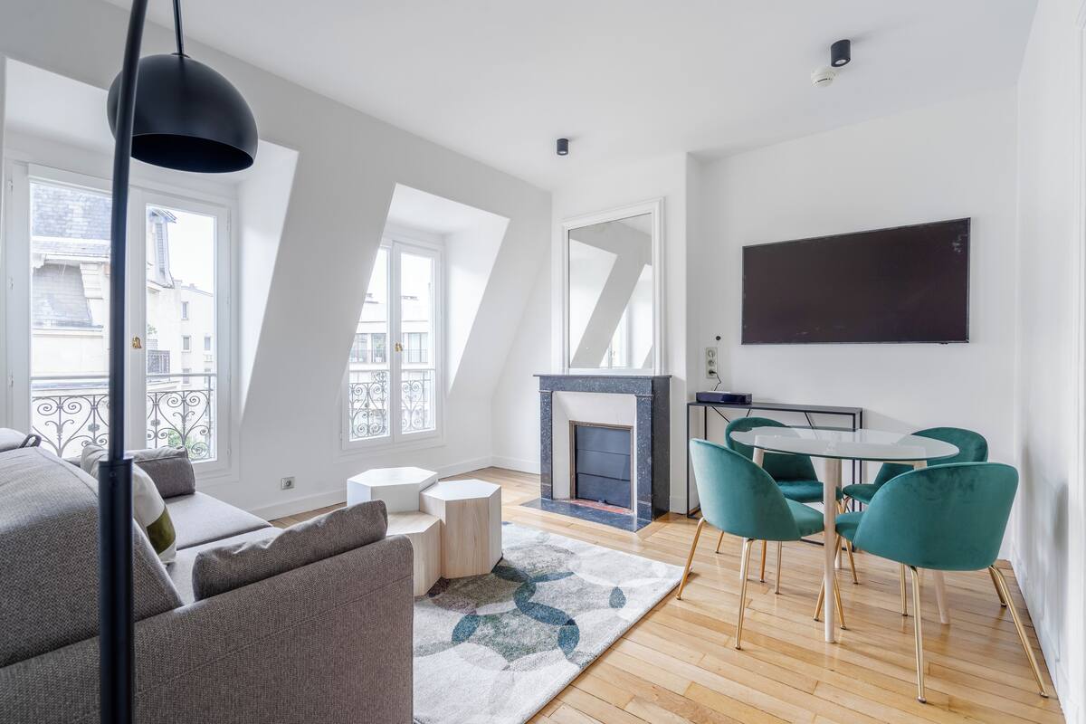 Ein modernes Wohnzimmer mit Kamin, einem Essbereich mit türkisfarbenen Stühlen und einem großen Fenster mit Blick auf Gebäude.