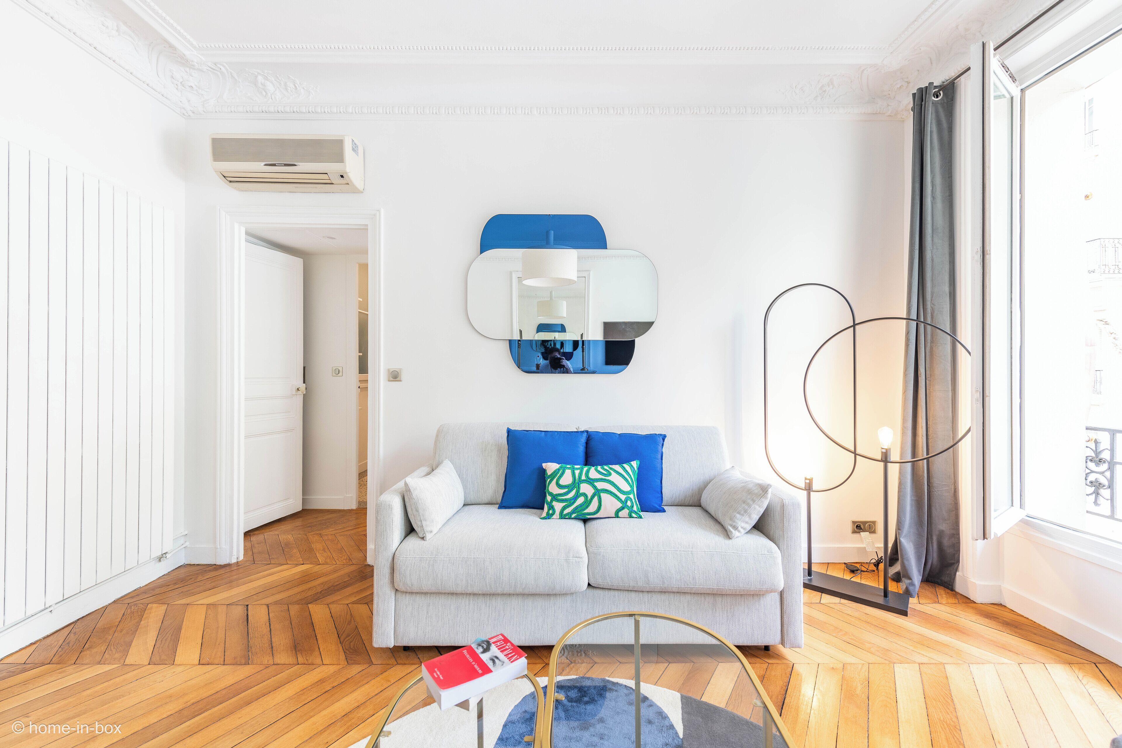 Un salon moderne avec un canapé gris clair, un coussin à motifs bleus et verts, un miroir rond et une lampe sur pied.