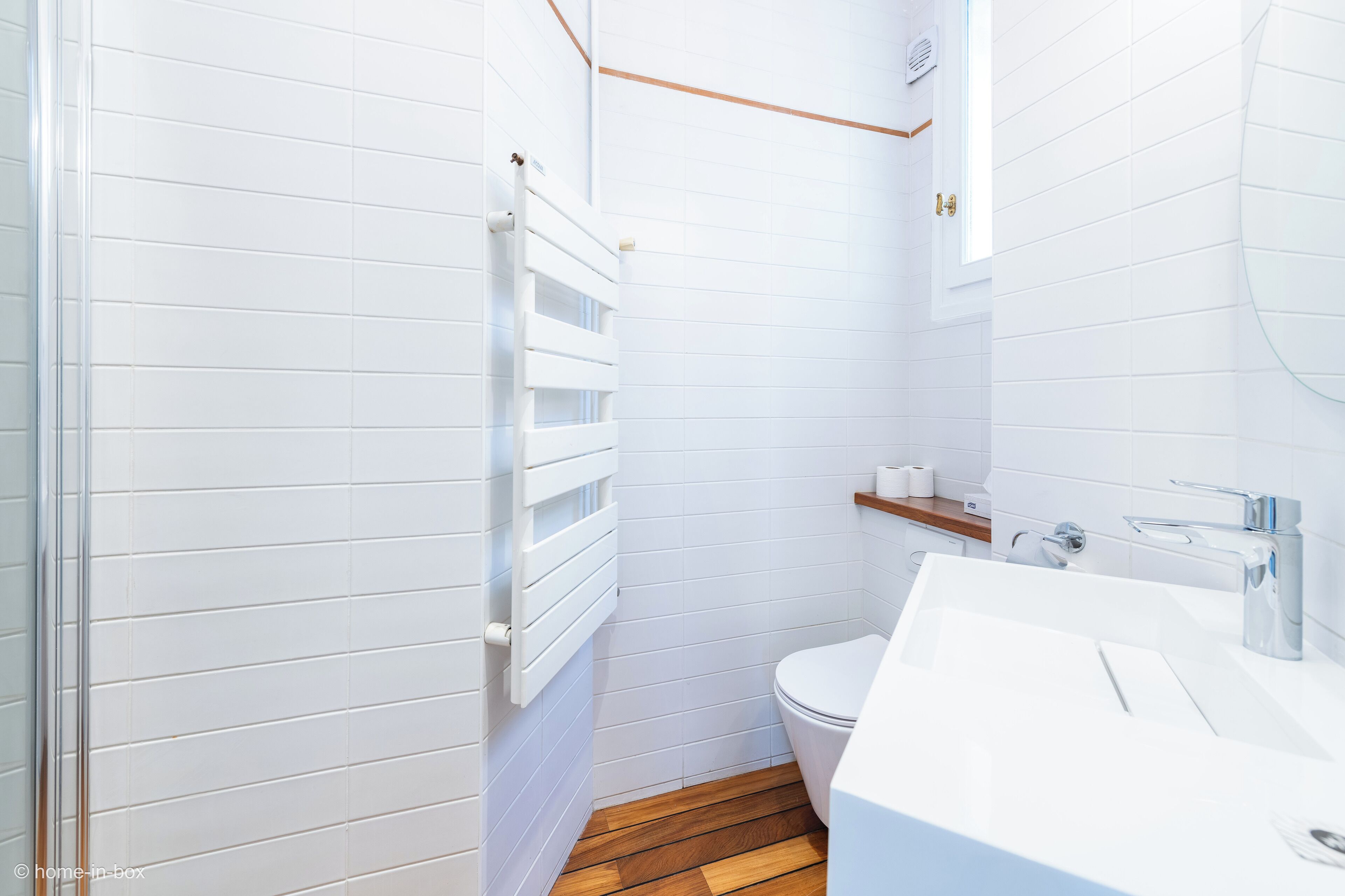 Un baño moderno con azulejos blancos, suelo de madera, inodoro blanco, lavabo con espejo y un perchero para toallas.