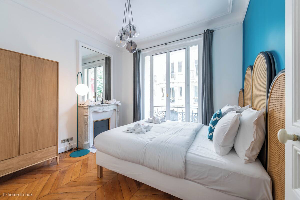 A bedroom with a large bed, a wooden wardrobe, a mirror, and a view of the city through large windows.
