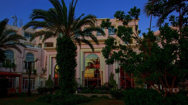A large, multi-story building with a pink facade, large windows, and a well-maintained garden with palm trees. - Regency Tunis Hotel (La Marsa)