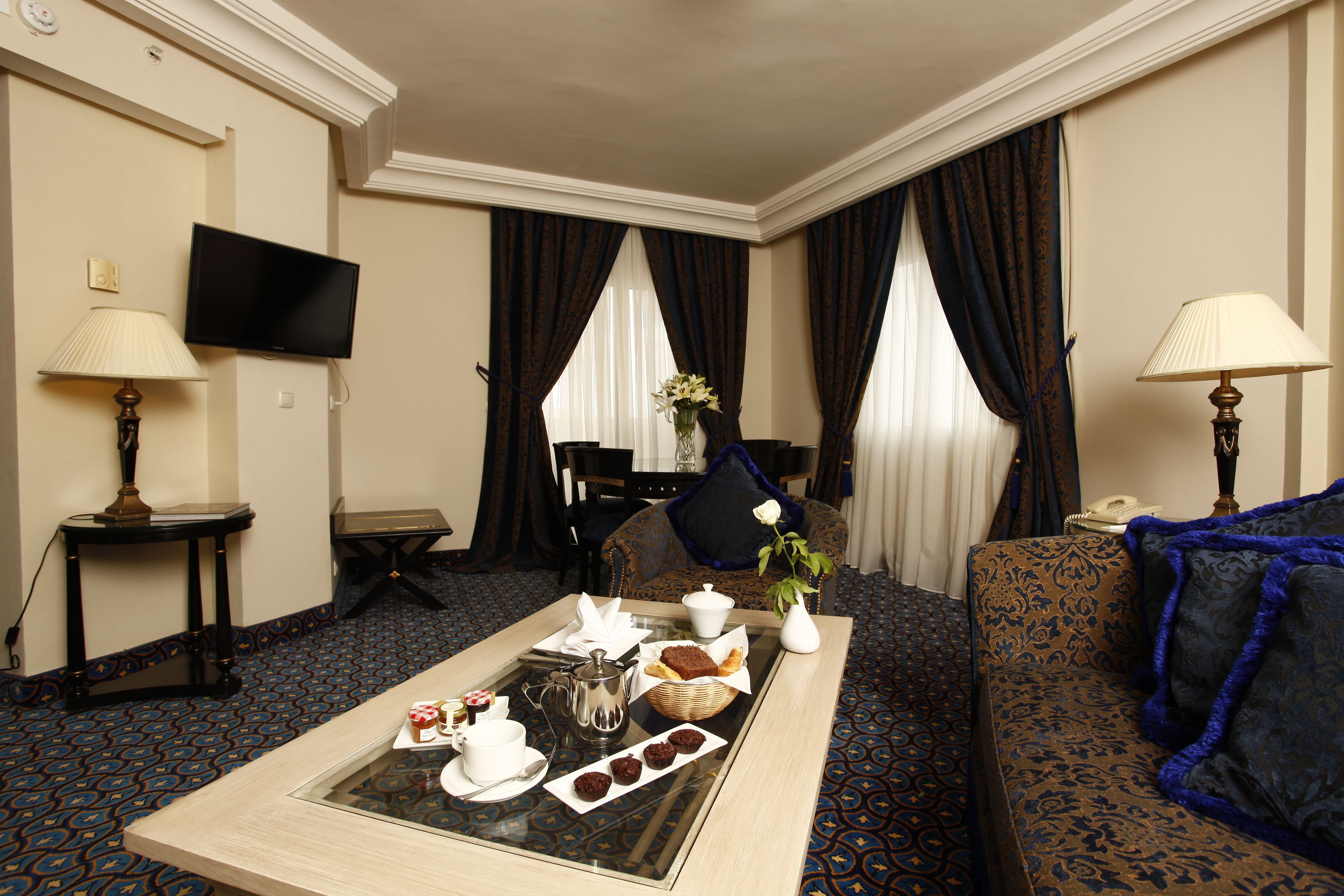 Une chambre d’hôtel avec une table à manger dressée, où l’on trouve du thé, des pâtisseries et un vase rempli de fleurs. Il y a également un canapé et un téléviseur fixé au mur.