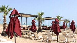 A beachside seating area with umbrellas and lounge chairs, a palm tree, and a clear view of the ocean. - Regency Tunis Hotel (La Marsa)