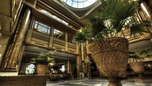 A spacious lobby with high ceilings, large windows, and ornate columns. There are several large potted plants and wicker furniture. - Regency Tunis Hotel (La Marsa)