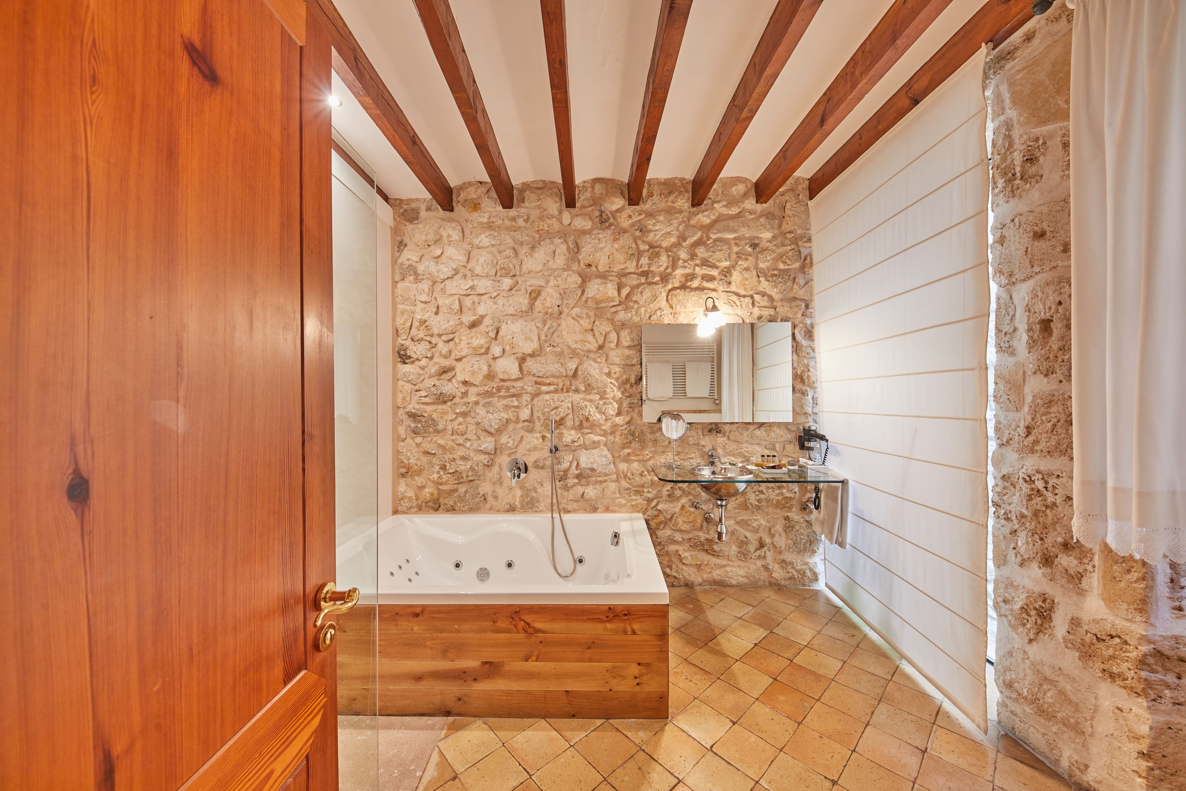 A bathroom with a stone wall, wooden beams, a bathtub, a glass sink, and a mirror.