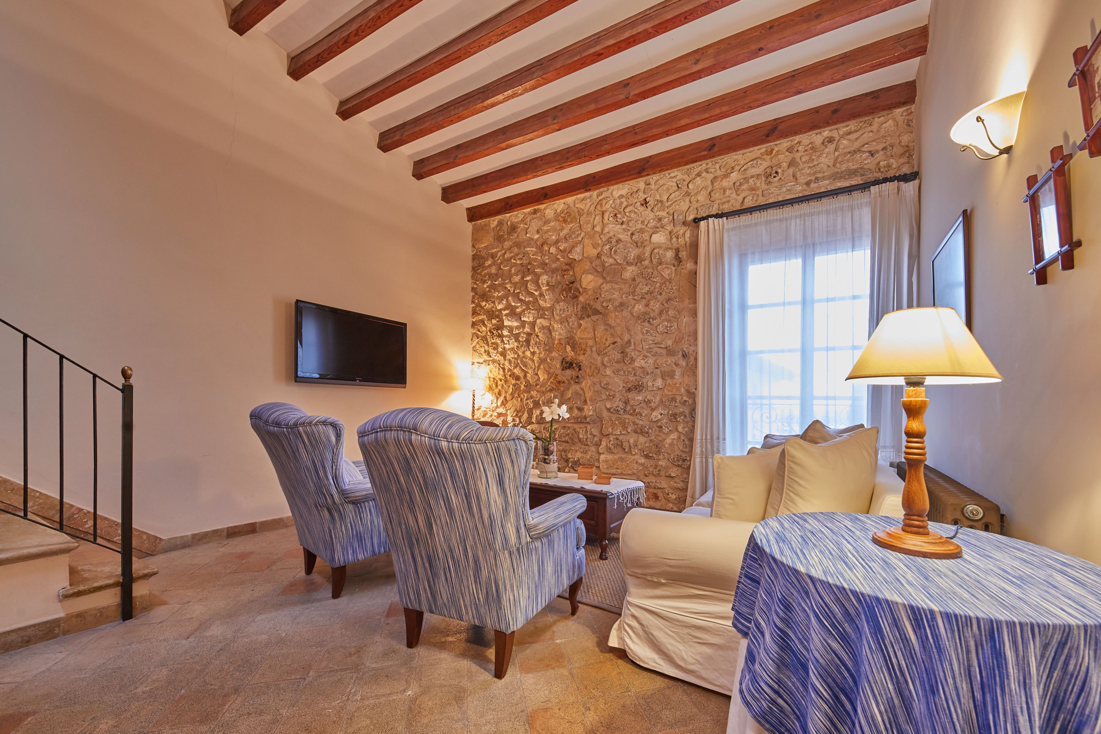 A living room with a stone wall, wooden beams, a TV, a sofa, two patterned armchairs, a table with a lamp, and a staircase.