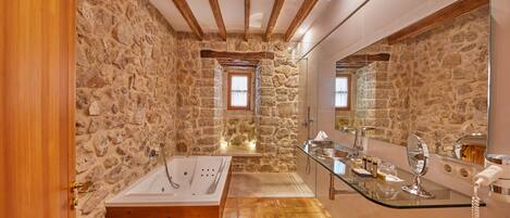Un baño con pared de piedra, vigas de madera, una bañera, tocador de cristal y un espejo grande.