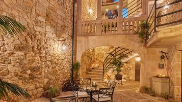 Un patio de piedra con una mesa y sillas de vidrio, rodeado de arcos y plantas.
