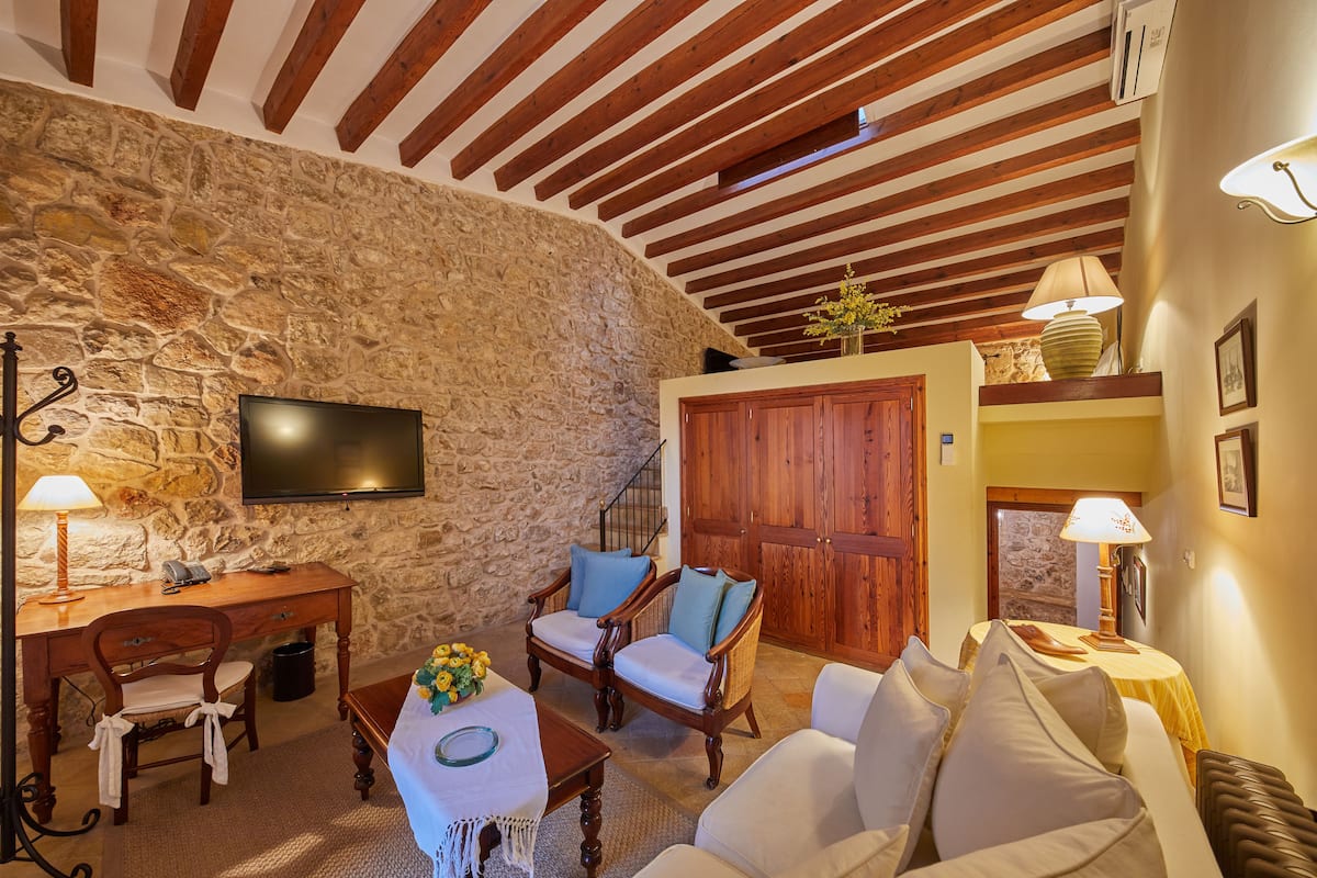 Una habitación acogedora con pared de piedra, vigas de madera en el techo, un televisor, un escritorio con lámpara, un sofá y una silla.