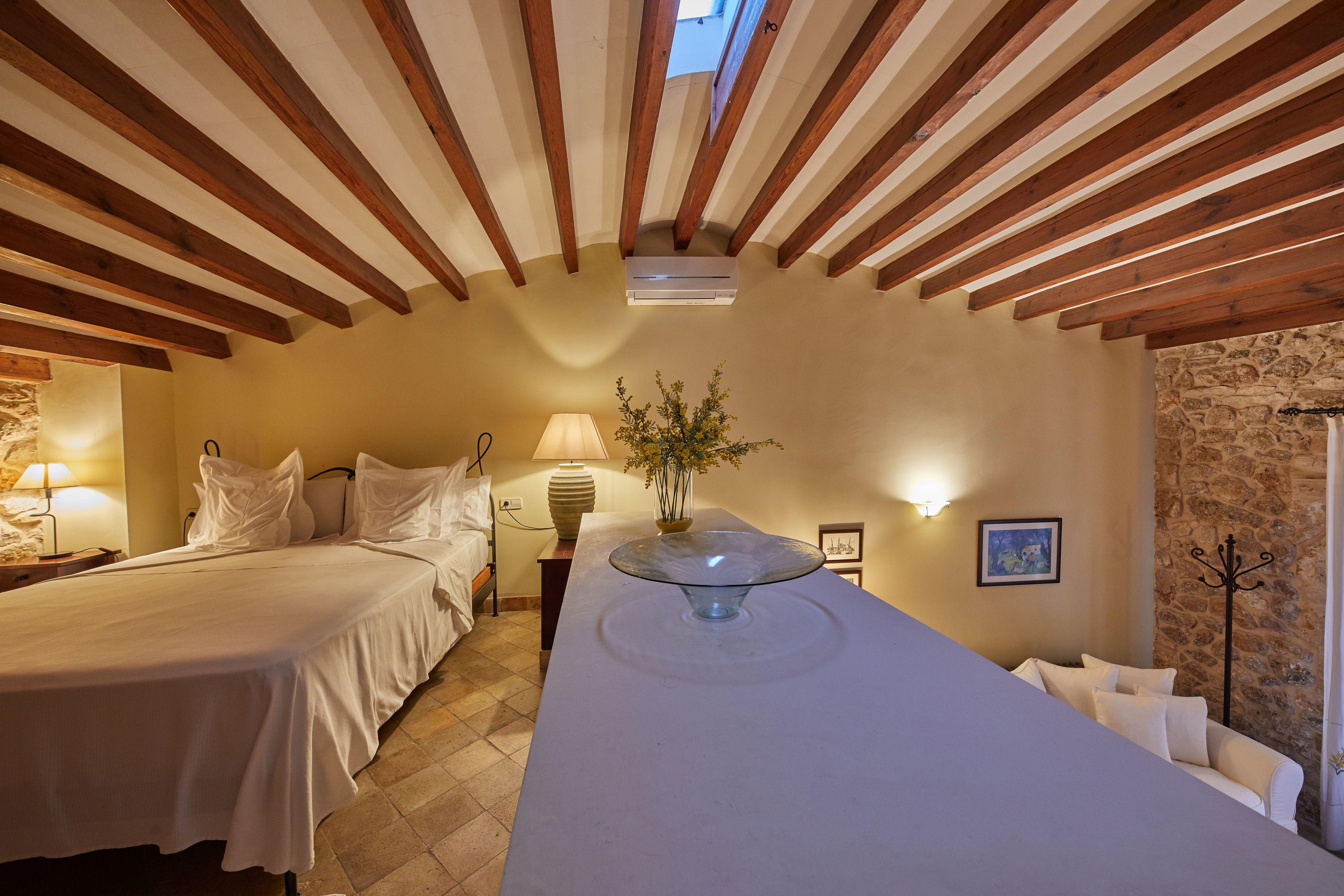 A hotel room with a large bed, a bedside table with a lamp, a glass-top table with a vase, and a stone wall.
