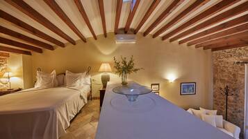 A hotel room with a large bed, a bedside table with a lamp, a glass-top table with a vase, and a stone wall.