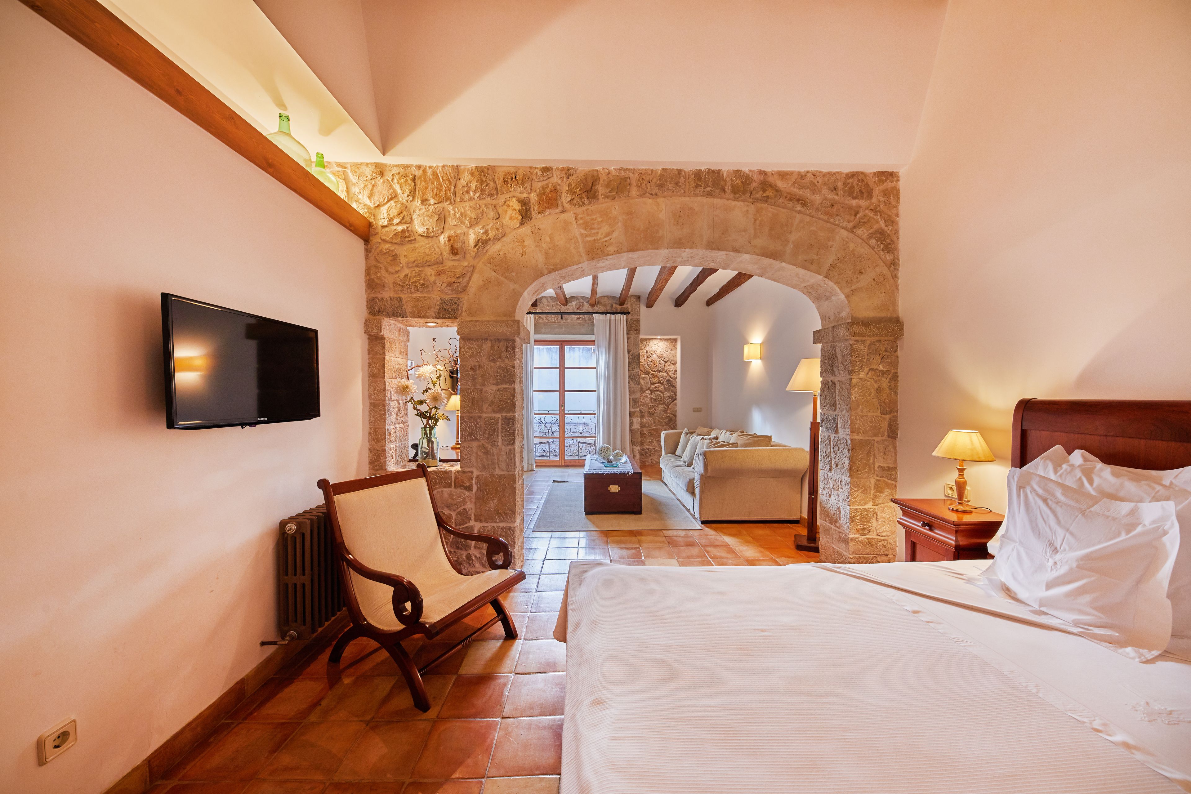 A bedroom with a stone archway, a bed, a chair, a TV, and a view of another room.