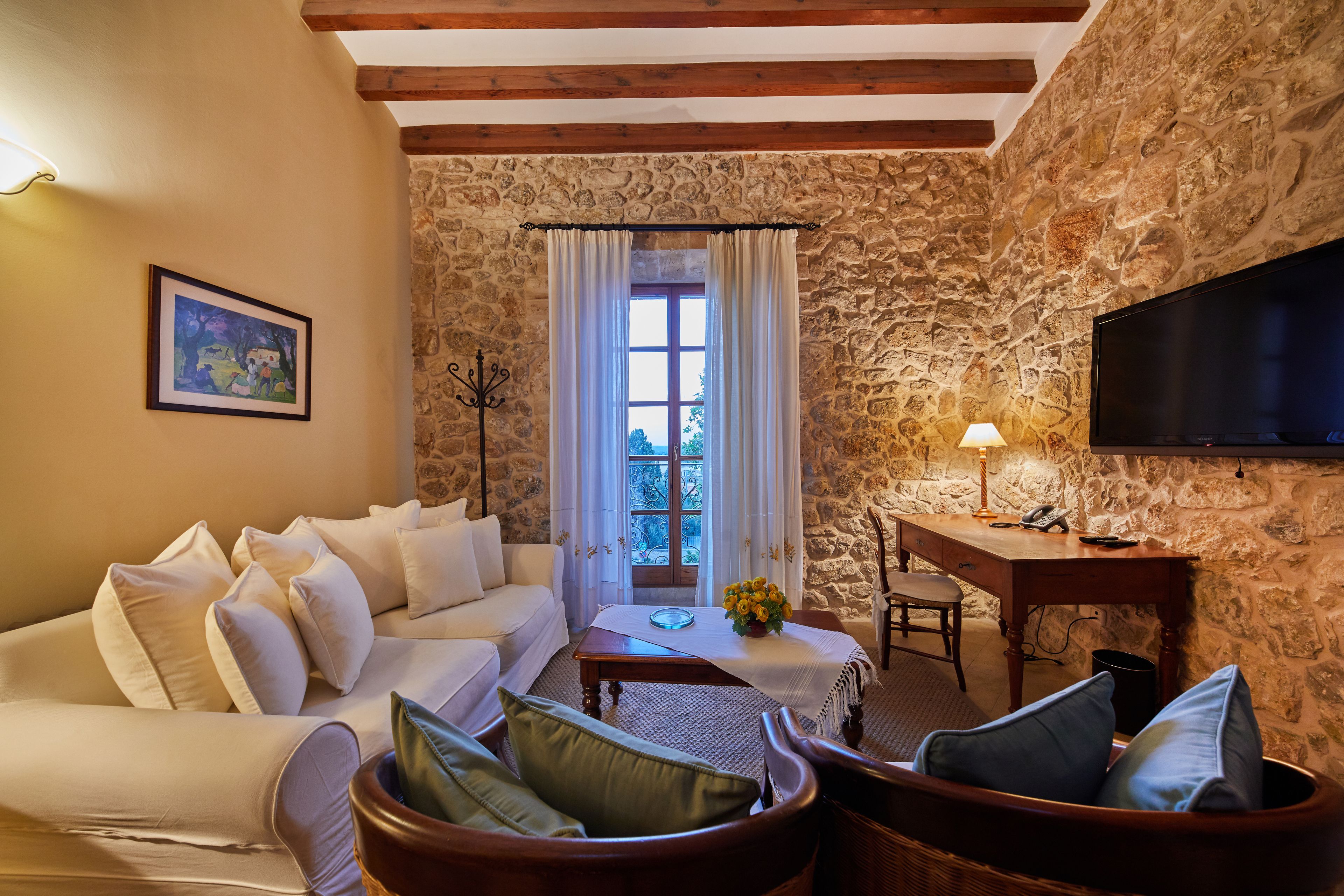 Un salon chaleureux avec un mur en pierre, un canapé blanc, une table basse en bois ornée d’un vase de fleurs, un téléviseur et un bureau avec une lampe.
