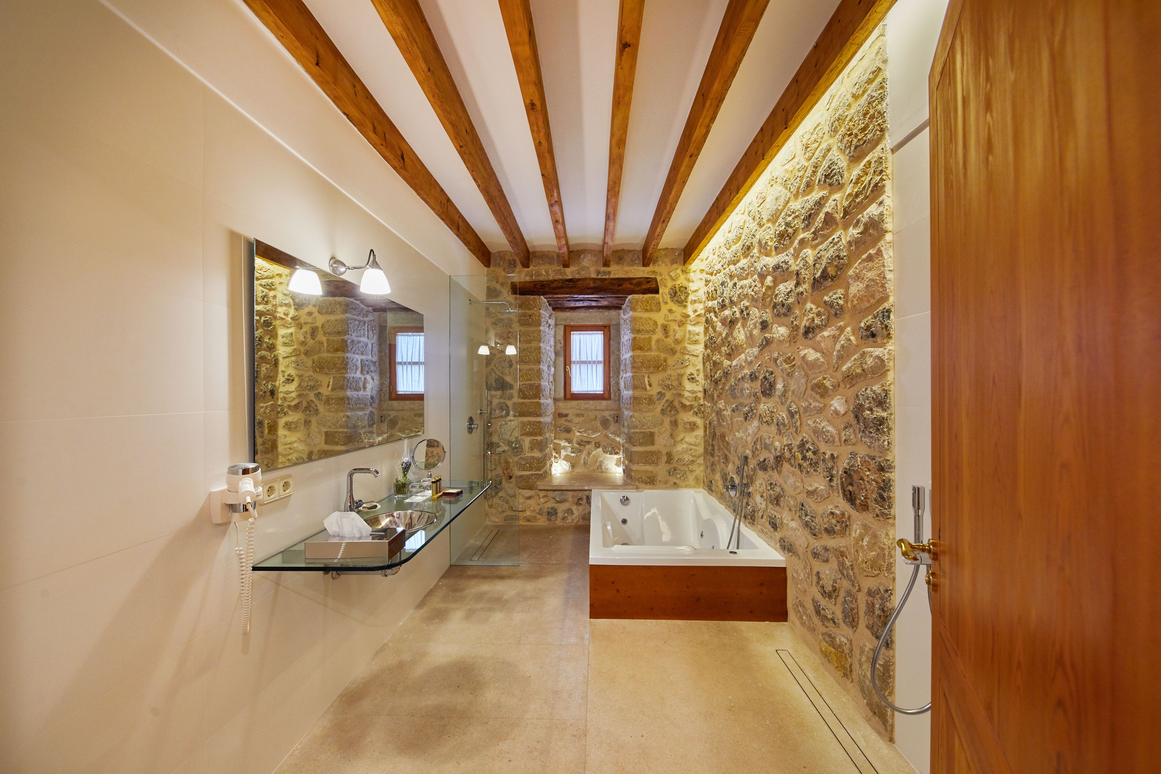 Un baño con pared de piedra, vigas de madera, cabina de ducha de vidrio, bañera de madera y tocador de madera con espejo.
