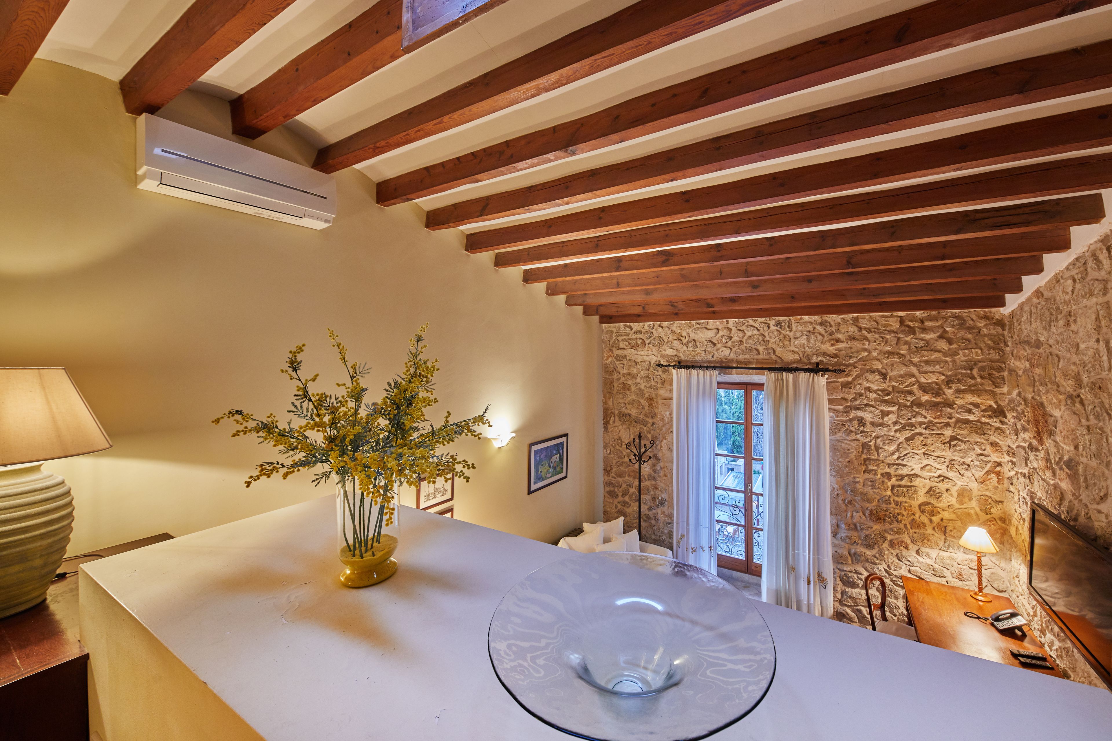 A room with a stone wall, wooden beams, a white ceiling, a glass table, a vase with yellow flowers, a lamp, and a window with curtains.