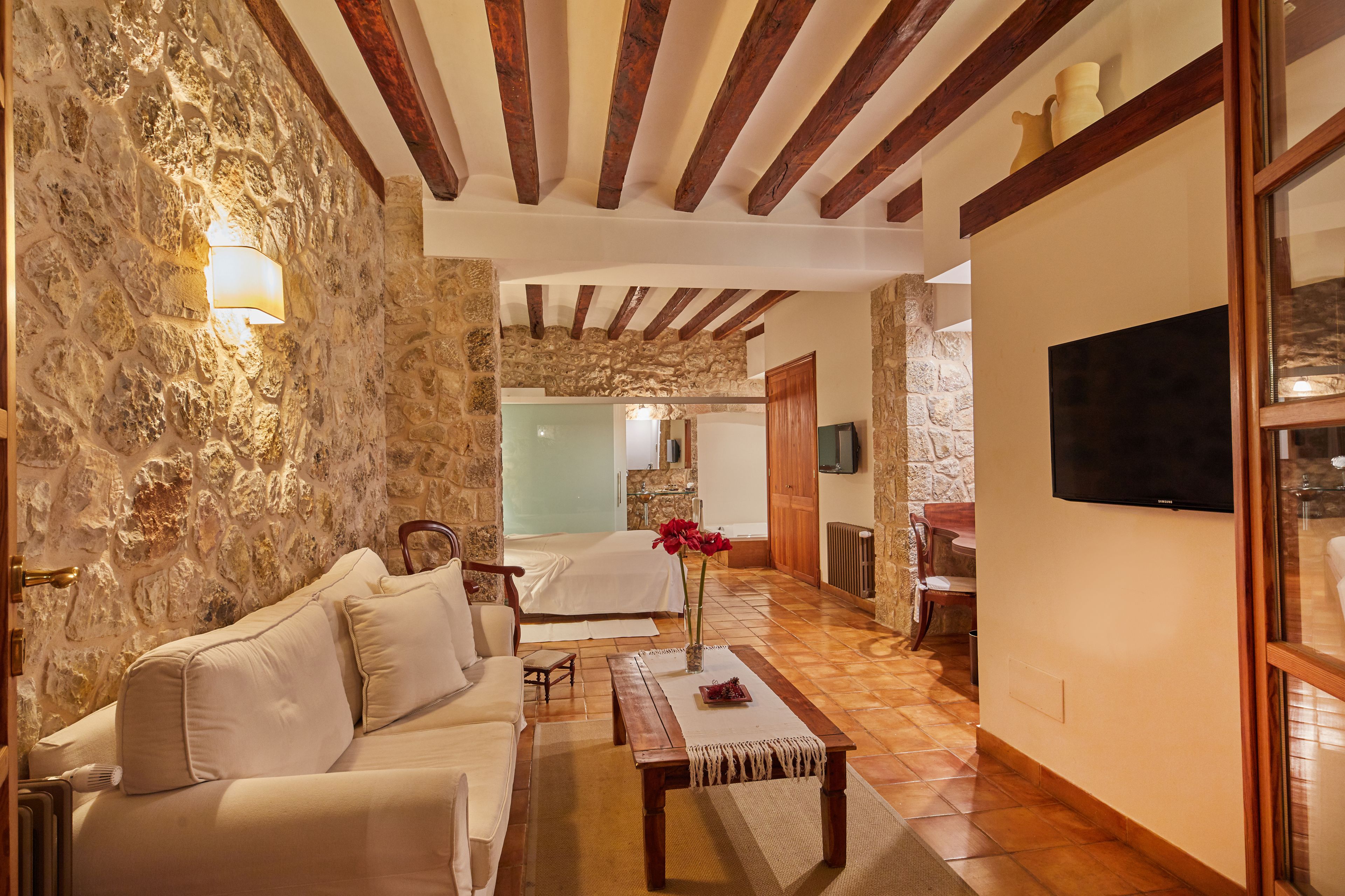 Una habitación de hotel acogedora con pared de piedra, techo de madera, cama, sofá, televisor y zona de comedor.
