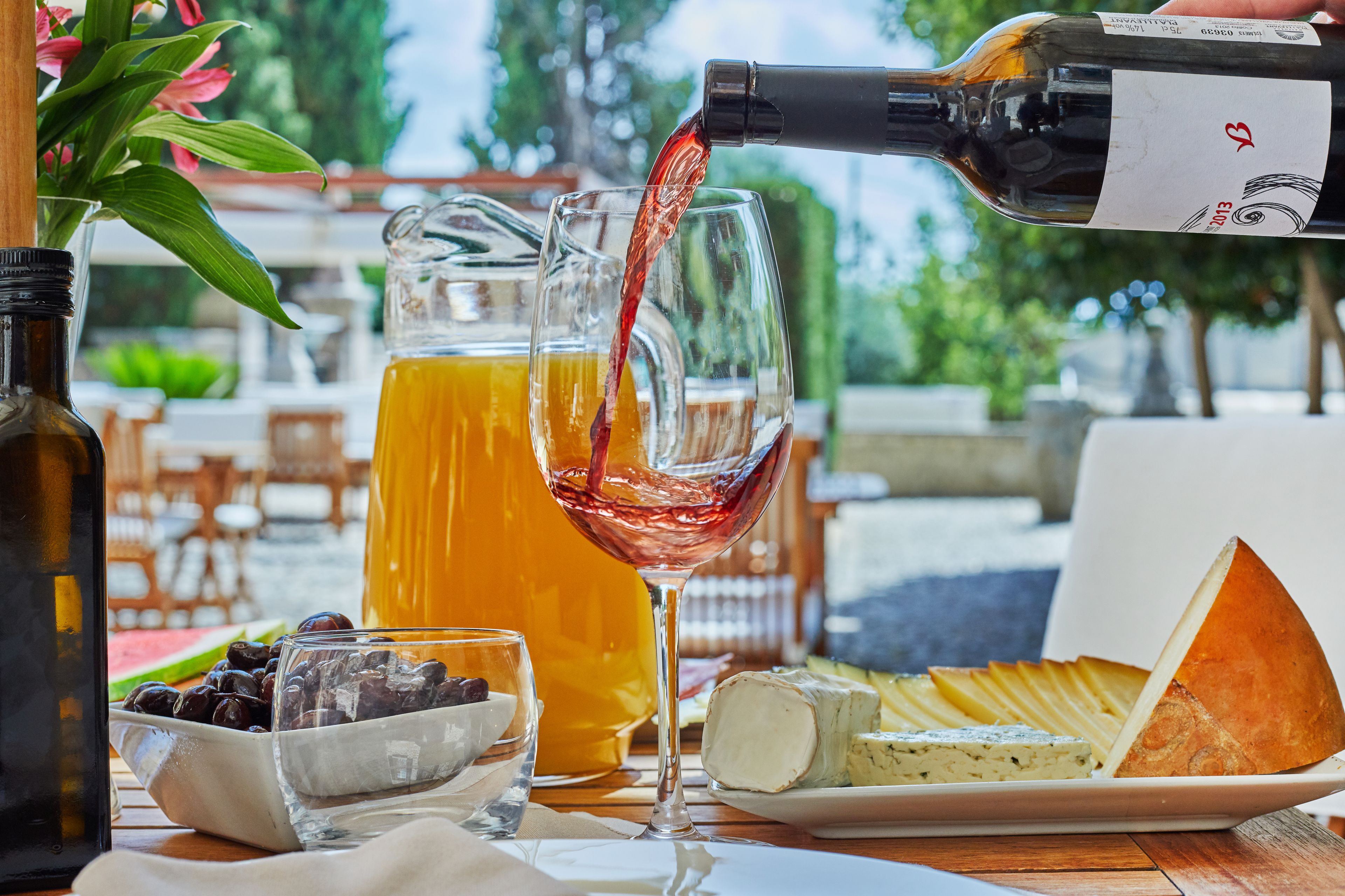 Una mesa con una botella de vino sirviéndose en una copa, una jarra de jugo de naranja, un cuenco de aceitunas, un plato de queso y una hogaza de pan.