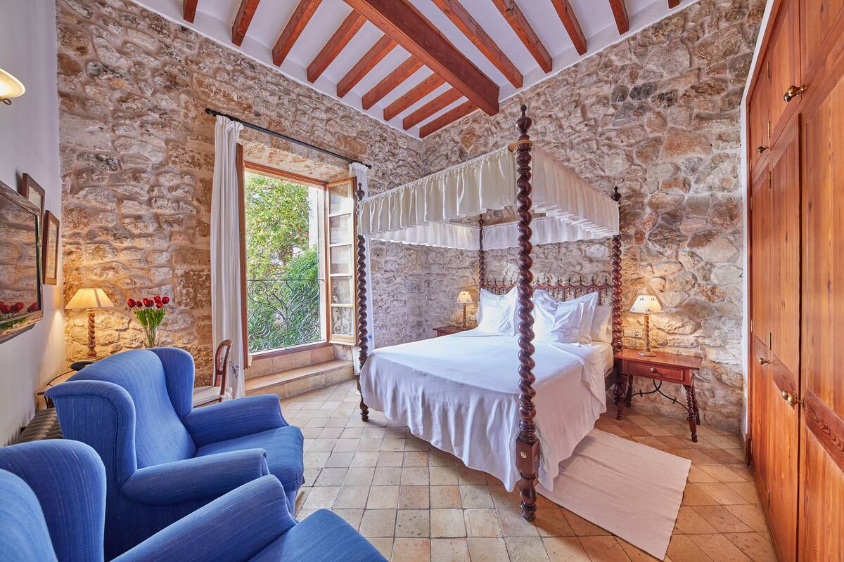 Un dormitorio con paredes de piedra, una cama con dosel, un armario de madera y un sillón azul.