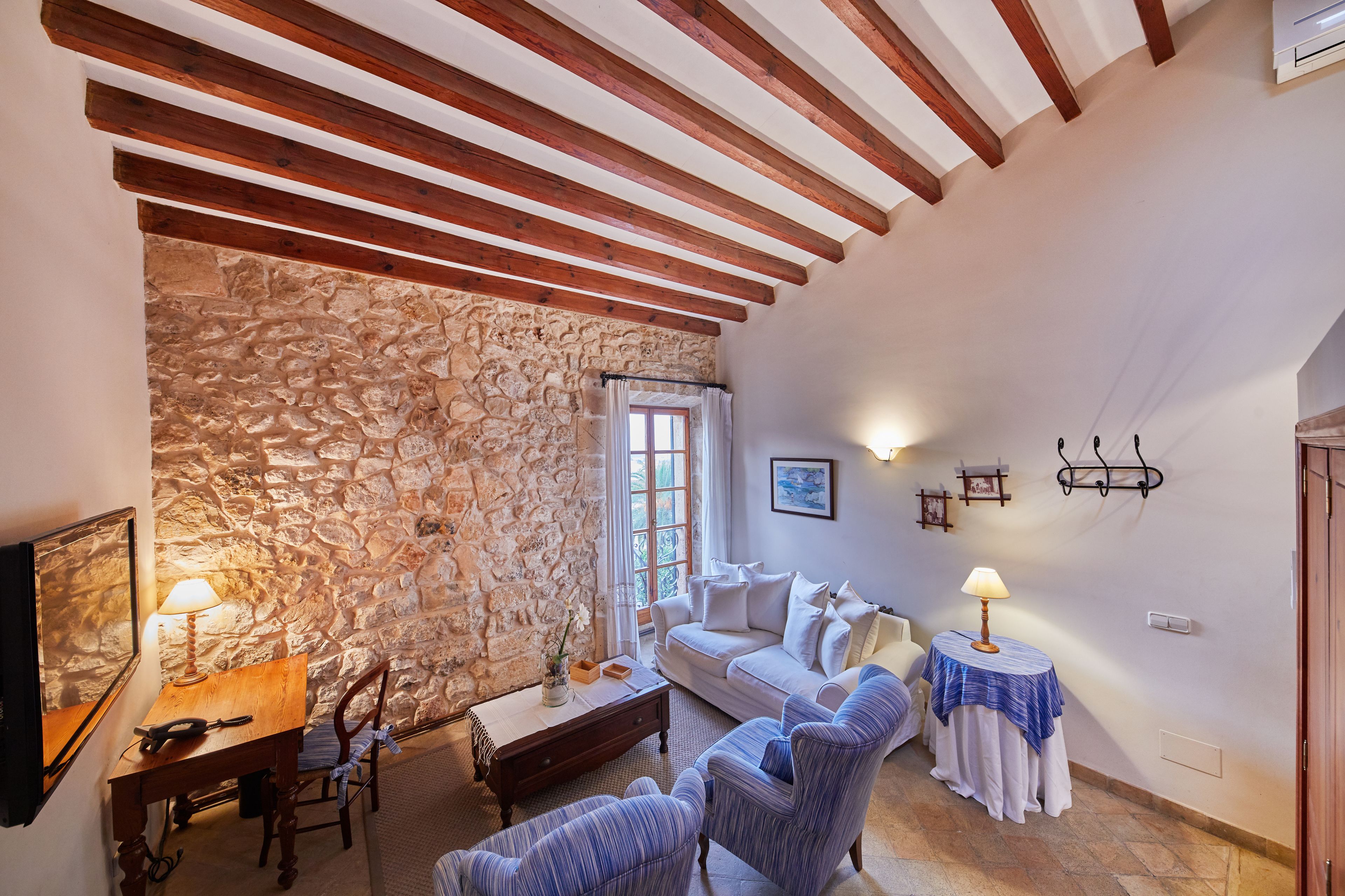 A cozy living room with a stone wall, wooden beams, a sofa, armchairs, a coffee table, and a small table with a lamp.