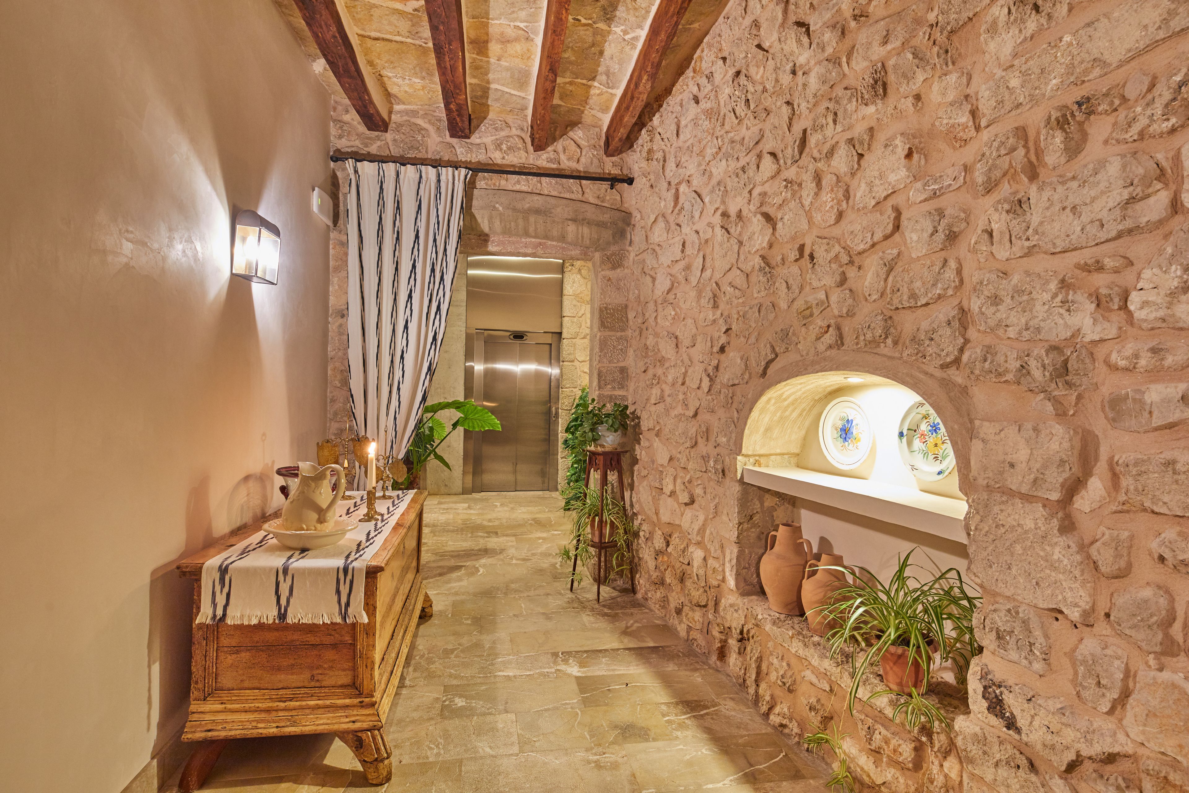 A narrow stone-walled corridor with a wooden bench, a wall-mounted light fixture, and a shelf with pottery.