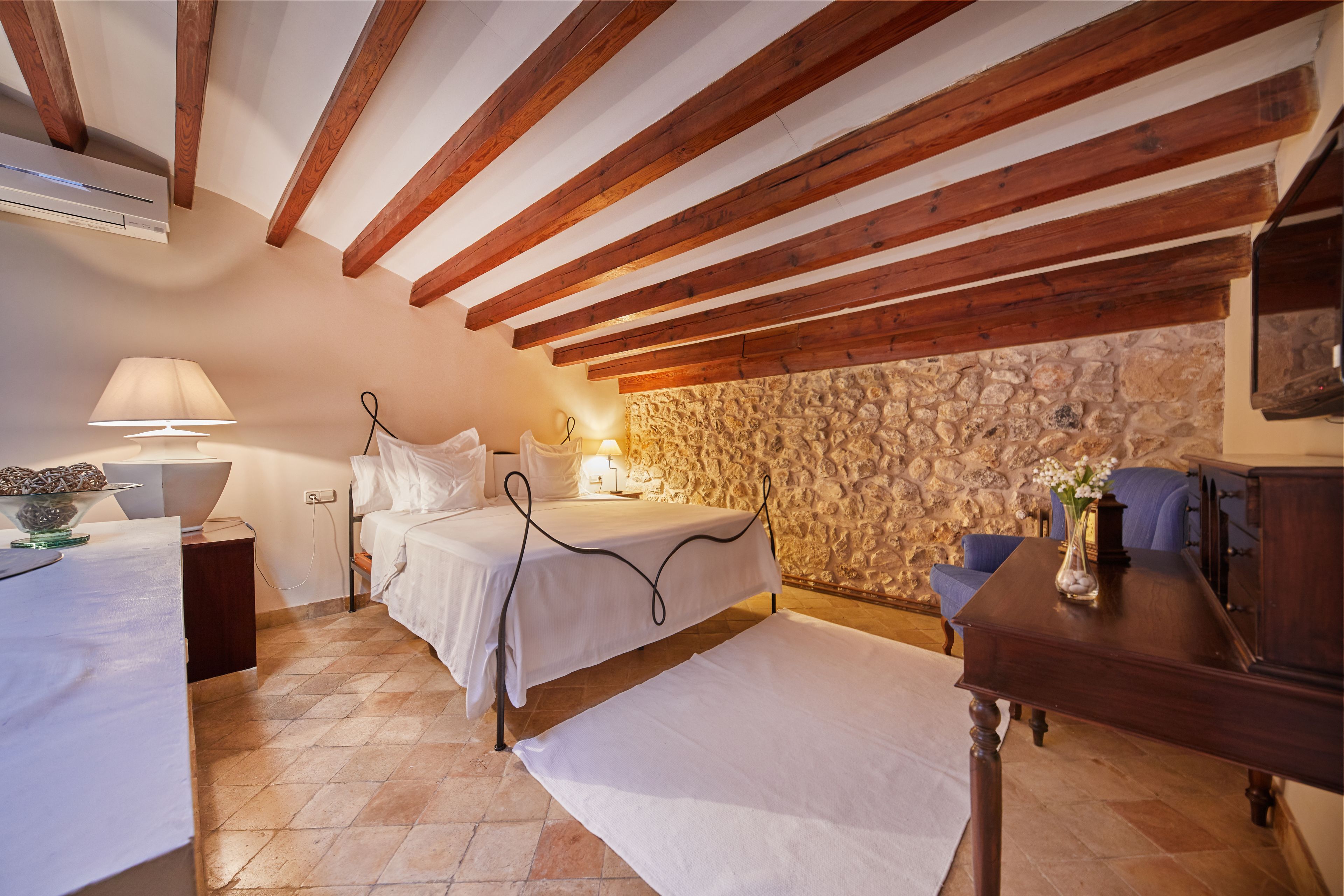 A bedroom with two beds, a stone wall, wooden beams, a stone fireplace, and a wooden nightstand with a lamp.