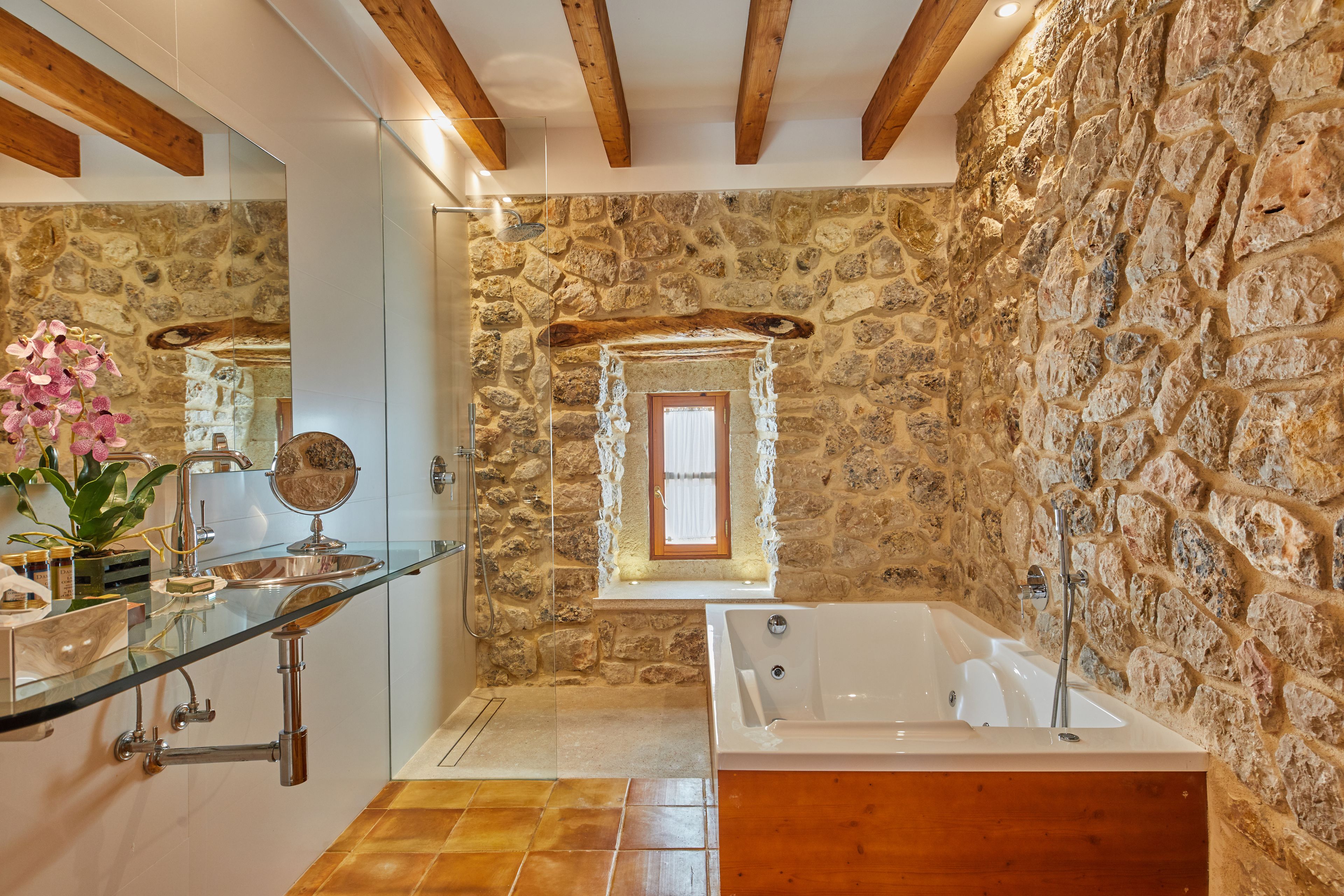 Une salle de bain avec un mur en pierre, un plafond en bois, une cabine de douche en verre, une grande baignoire et un miroir.