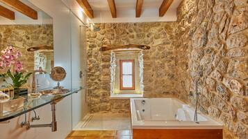 Une salle de bain avec un mur en pierre, un plafond en bois, une cabine de douche en verre, une grande baignoire et un miroir.