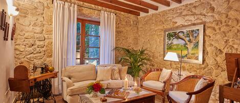 A cozy living room with a stone wall, wooden beams, a sofa, chairs, a coffee table, and a sewing machine.