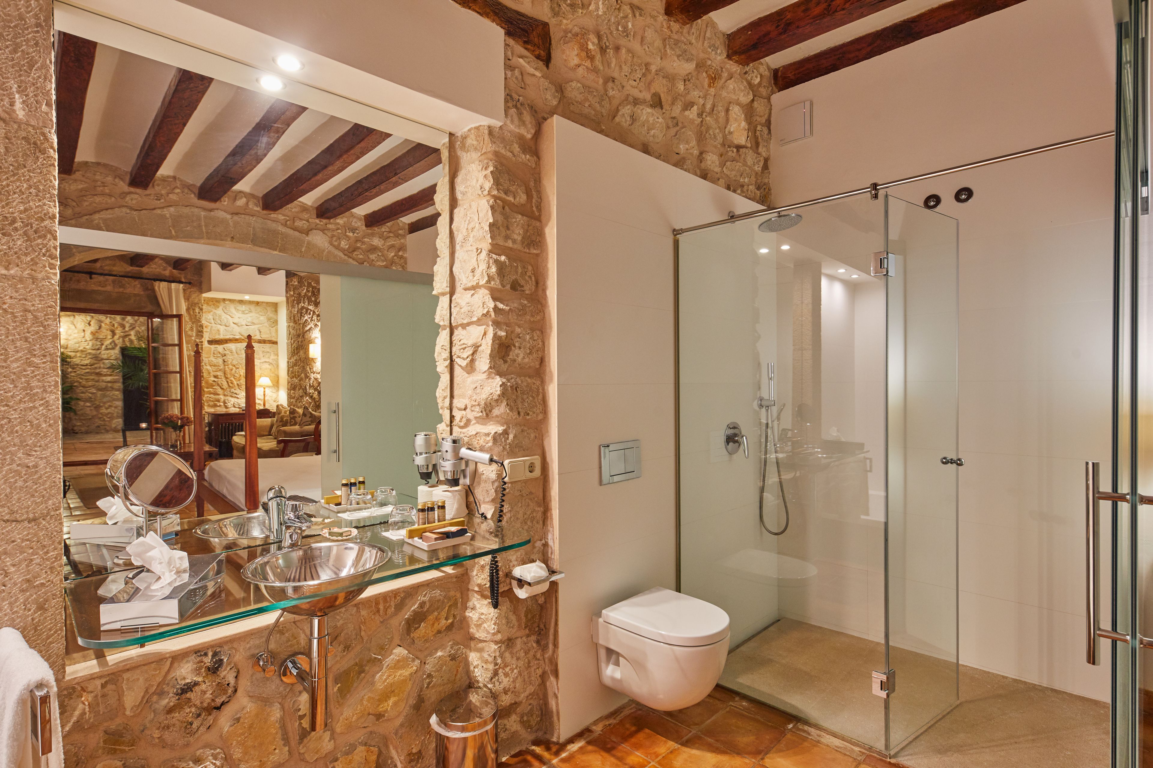 Une salle de bain avec un mur en pierre, une douche en verre, des toilettes blanches et un lavabo en verre avec un miroir.