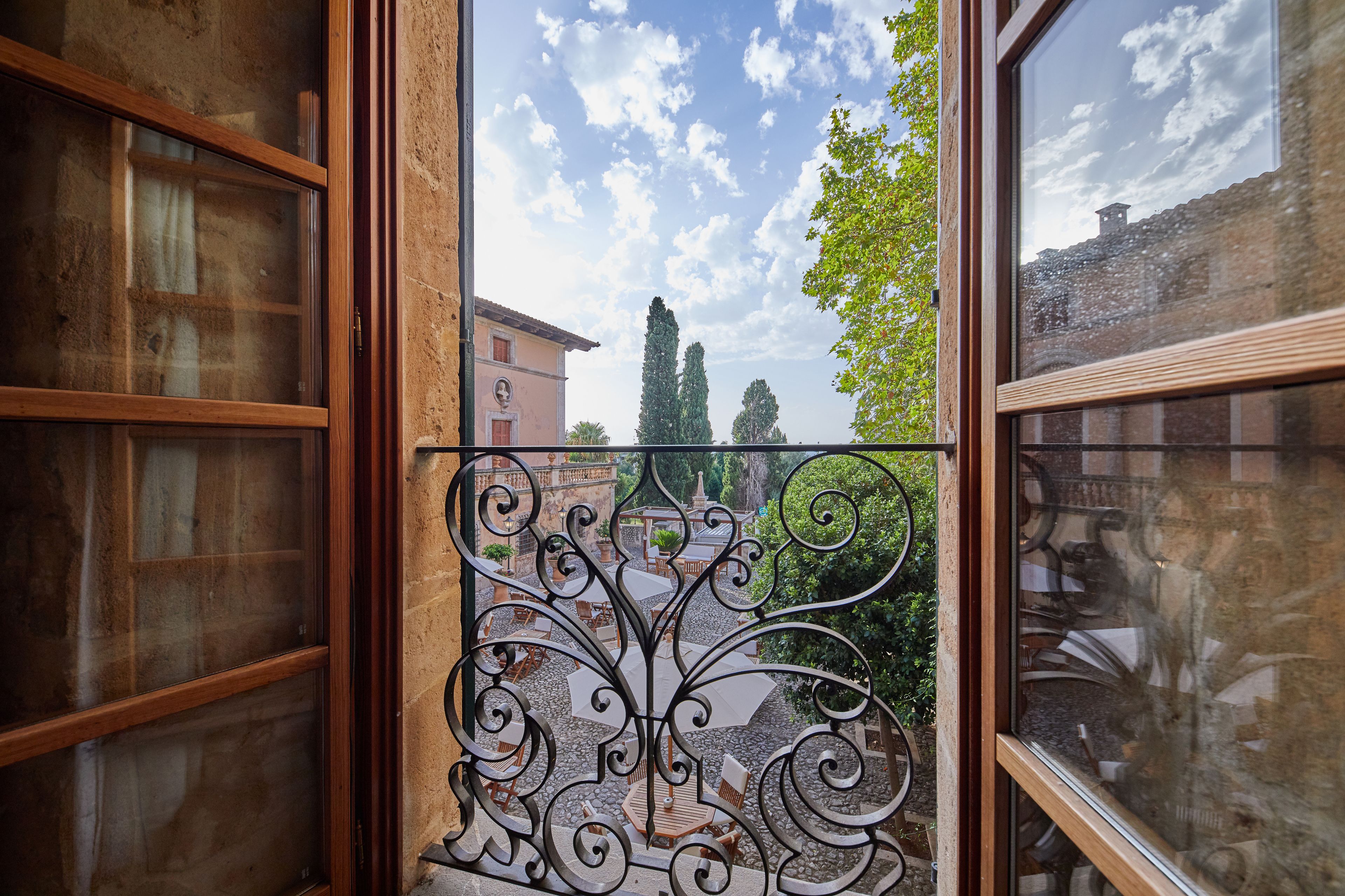 A window with a decorative iron grille, a view of a courtyard with outdoor furniture, and a building in the background.
