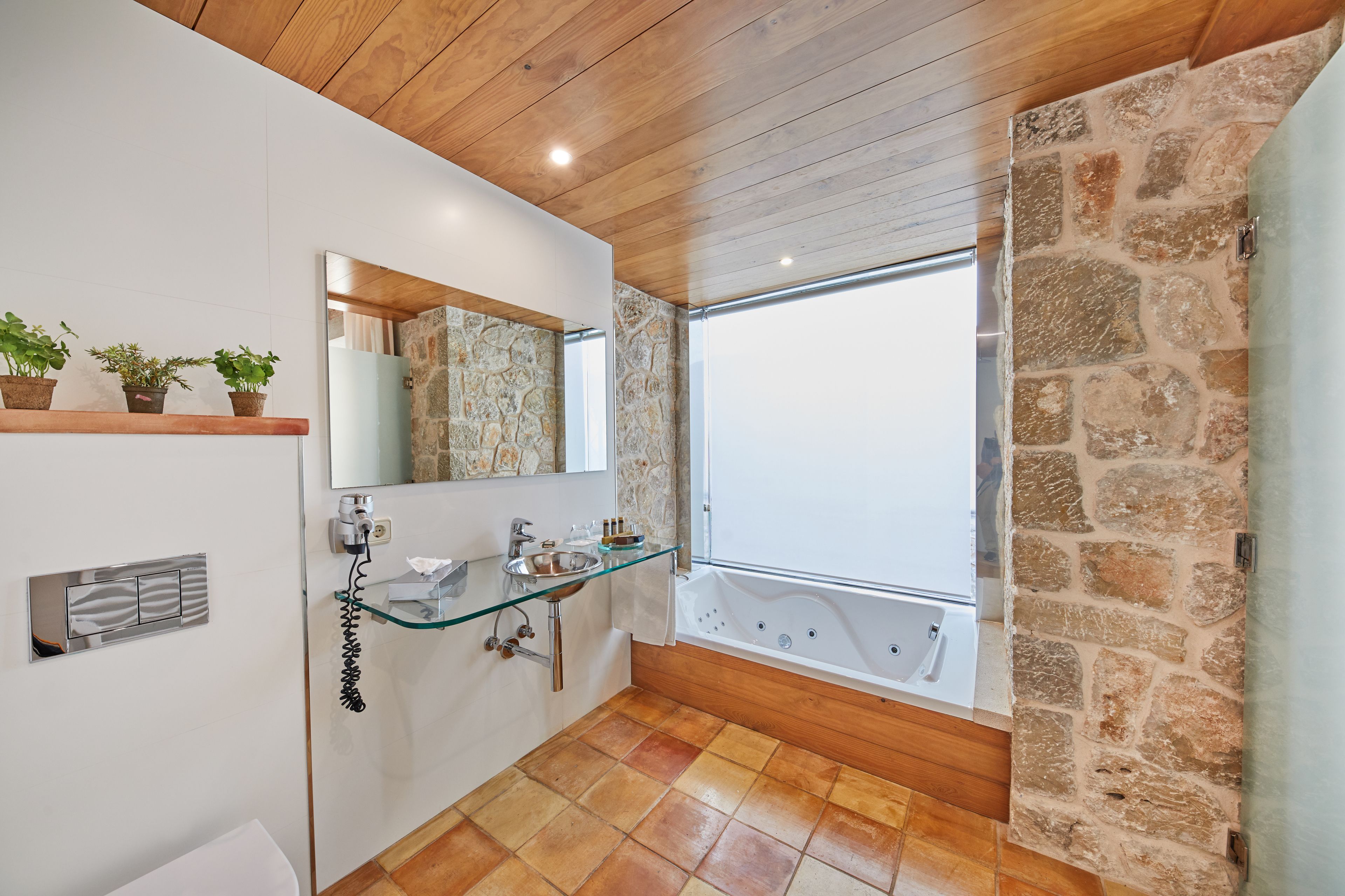Un baño moderno con un lavabo de vidrio, dos lavabos, una bañera grande y una pared de piedra.
