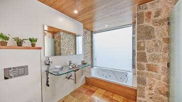 Un baño moderno con un lavabo de vidrio, dos lavabos, una bañera grande y una pared de piedra.