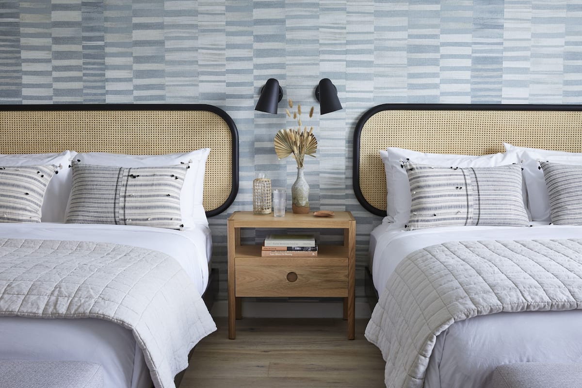 A bedroom with two beds, a nightstand with books and a vase, and two wall-mounted lamps.